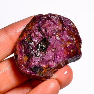 Puede incluir: Una gema de rubí en bruto, sin cortar, con un tono rojo-púrpura intenso. La superficie es rugosa y texturizada, con áreas más oscuras y toques de naranja. Un hermoso ejemplo de especímenes minerales en bruto.