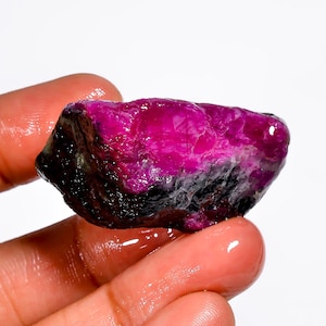 May include: A raw ruby gemstone, displaying a rich magenta colour. The uncut mineral shows a mix of deep magenta and black hues, with a slightly textured surface. The gemstone is held in a hand against a white background.