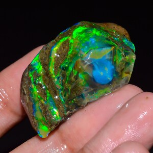 May include: A vibrant, raw opal gemstone with a striking display of color. The stone features a mix of green, blue, and brown hues, with a central blue area. The opal is held in a hand, showcasing its natural, rough texture.