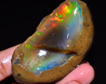 May include: A raw, unpolished opal gemstone featuring a smooth, translucent center with vibrant flashes of red, green, and blue fire. The stone is encased in a rough, textured, earthy brown rock matrix with visible pores and natural surface cracks.