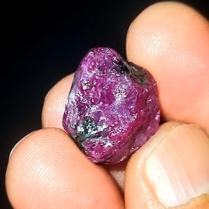 Peut inclure: Une pierre précieuse de rubis rouge foncé avec une finition brute et naturelle. Le rubis a une couleur rouge foncé avec des nuances de violet et de noir. La pierre précieuse est tenue dans une main.