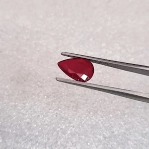 以下が含まれることがあります： ピンセットで保持された、洋梨型の濃い赤色の宝石。宝石はルビーです。