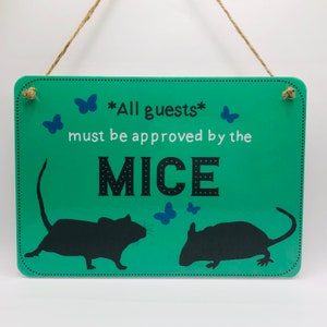 May include: A green sign with the text "*All guests* must be approved by the MICE" in black and white lettering. Two black silhouettes of mice are on either side of the text. There are blue butterfly silhouettes scattered around the sign.