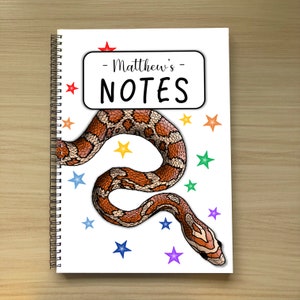 May include: A white notebook with a spiral binding. The cover features a black and white illustration of a snake with a red star on top. The text "Matthew's NOTES" is written in black on a white rectangle.