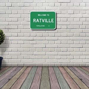 May include: A green road sign with white text that reads "Welcome to Ratville Population 3". The sign is mounted on a white brick wall. A small green bush is in a blue pot on the left side of the image.