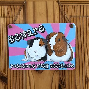 May include: A pink and blue sign with the text "Beware Potatoes with attitude" and a picture of two guinea pigs. One guinea pig is brown and white, and the other is black and white.