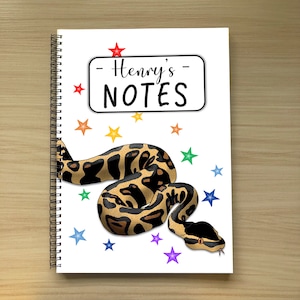 May include: A white spiral-bound notebook with a snake illustration. The notebook cover has the text "Henry's NOTES" in a black font, with colorful star shapes scattered around the design.