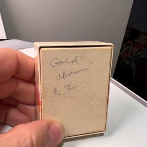 May include: A rectangular, off-white cardboard jewelry box with a lid. The top surface of the lid features handwritten cursive text in dark ink that reads Gold chain and the number 2, with a small symbol resembling a dollar sign above the number.