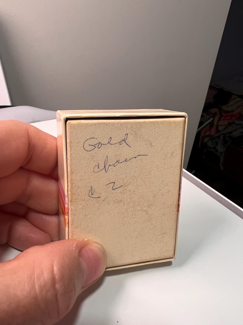 May include: A rectangular, off-white cardboard jewelry box with a lid. The top surface of the lid features handwritten cursive text in dark ink that reads Gold chain and the number 2, with a small symbol resembling a dollar sign above the number.