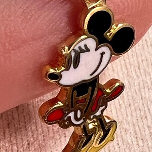 May include: A gold-toned metal charm featuring a stylized enamel character with black ears, a white face, and a red outfit. The charm has a small circular loop at the top for attachment, with clear outlines defining the character's features and clothing.
