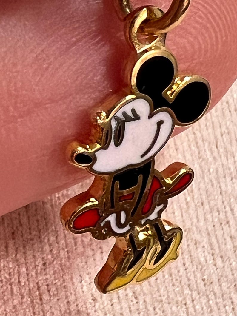 May include: A gold-toned metal charm featuring a stylized enamel character with black ears, a white face, and a red outfit. The charm has a small circular loop at the top for attachment, with clear outlines defining the character's features and clothing.