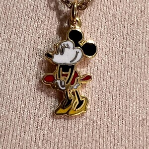 May include: A gold-toned metal necklace featuring a pendant of a cartoon mouse character. The character has a white face, black ears, a red skirt, and yellow shoes, with black outlines defining the figure. The pendant hangs from a gold chain.