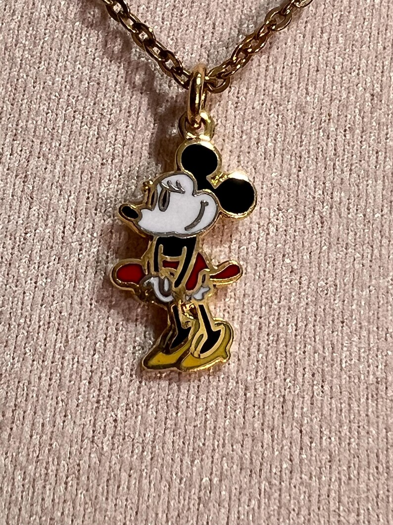 May include: A gold-toned metal necklace featuring a pendant of a cartoon mouse character. The character has a white face, black ears, a red skirt, and yellow shoes, with black outlines defining the figure. The pendant hangs from a gold chain.
