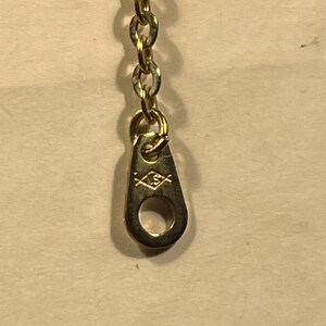 May include: A gold-toned metal chain necklace featuring a teardrop-shaped tag at the end. The tag displays a small, engraved diamond-shaped hallmark containing the letters LS, positioned below the circular link of the chain.