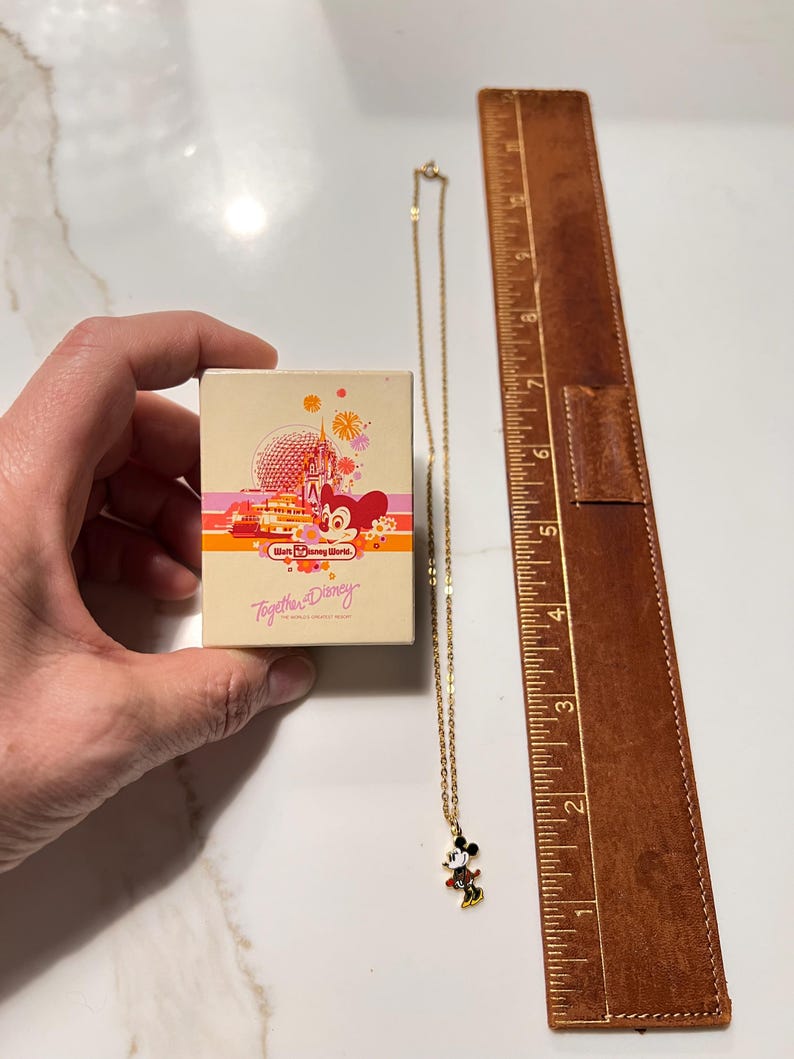 May include: A gold-tone chain necklace with a Mickey Mouse pendant, accompanied by a rectangular Walt Disney World box featuring a graphic of Spaceship Earth and the text Together at Disney. A brown ruler shows the necklace is 9 inches long.