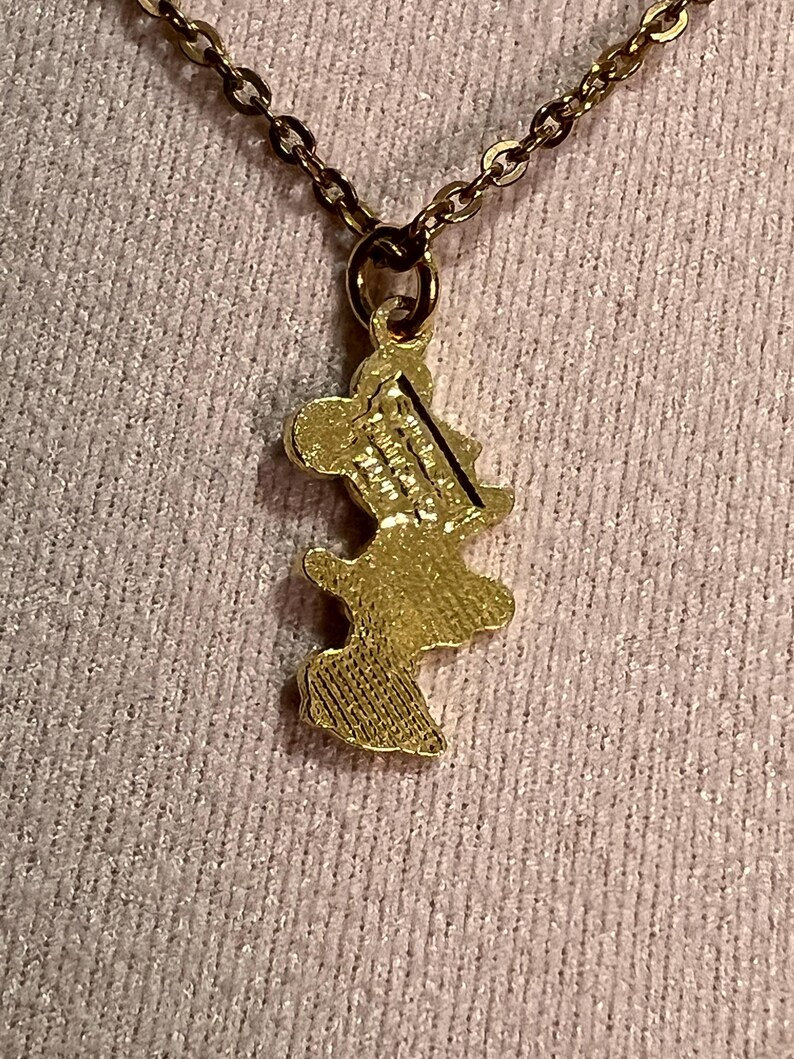 May include: A gold-toned metal pendant shaped like a cartoon mouse silhouette, suspended from a matching gold-toned link chain. The back of the pendant features embossed text that reads Disney and China, with a textured finish on the lower portion.