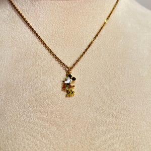 May include: A gold-toned metal chain necklace featuring a small, colorful enamel pendant of a cartoon mouse character. The pendant depicts the character in black, white, red, and yellow colors, suspended from a delicate link chain.