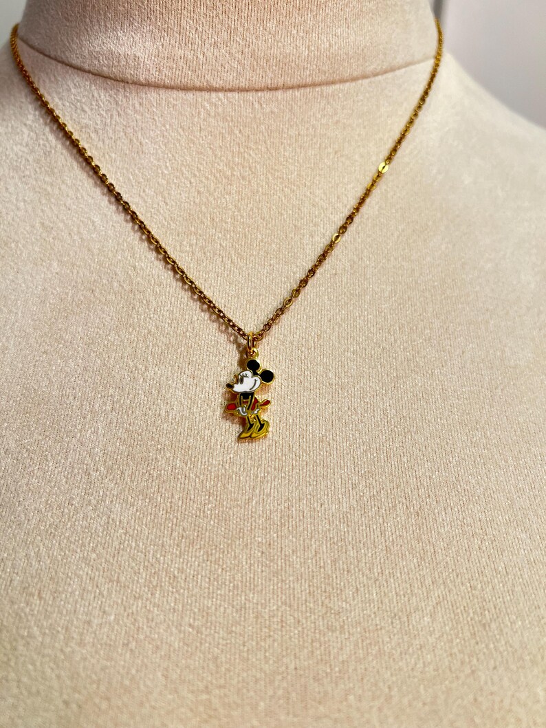 May include: A gold-toned metal chain necklace featuring a small, colorful enamel pendant of a cartoon mouse character. The pendant depicts the character in black, white, red, and yellow colors, suspended from a delicate link chain.