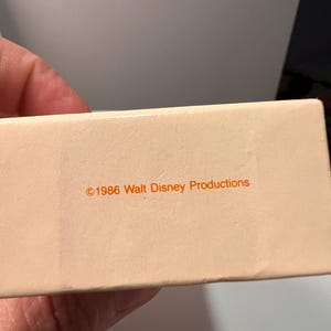 May include: A rectangular, off-white cardboard box featuring the orange printed text &copy;1986 Walt Disney Productions centered on the side. The surface of the box is smooth and matte, displaying clear, legible typography in a simple sans-serif font.