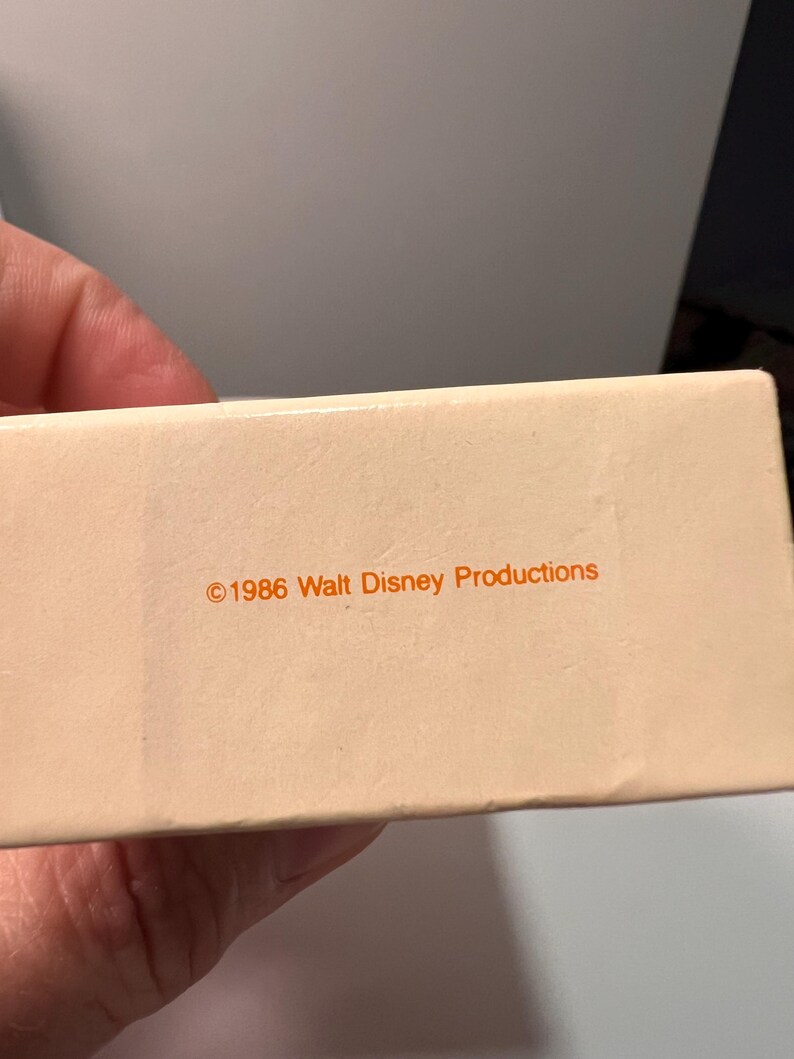 May include: A rectangular, off-white cardboard box featuring the orange printed text &copy;1986 Walt Disney Productions centered on the side. The surface of the box is smooth and matte, displaying clear, legible typography in a simple sans-serif font.