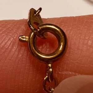 May include: A gold-toned spring ring clasp attached to a delicate chain. The circular clasp features a small lever mechanism on the side for opening and closing, with a small metal tag attached to the jump ring connecting it to the chain links.