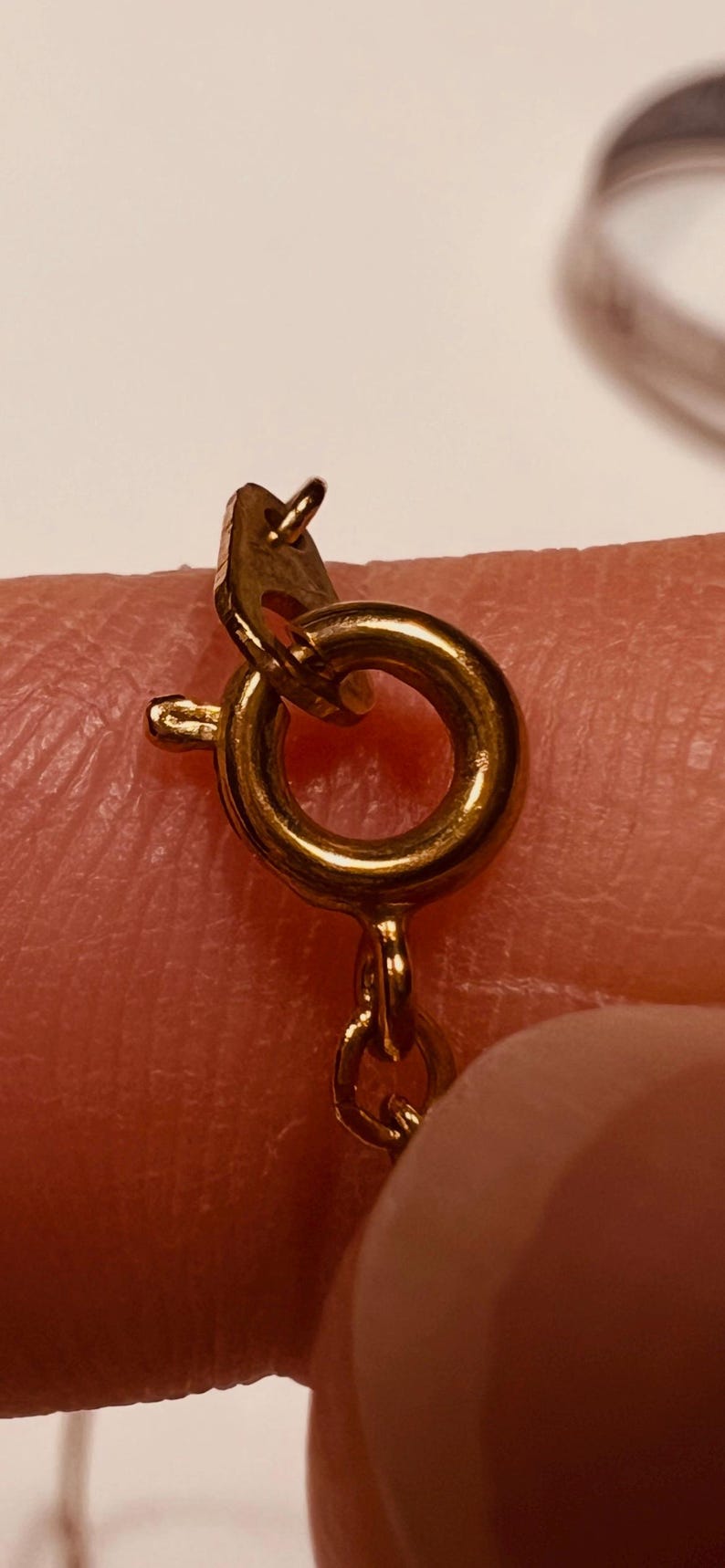 May include: A gold-toned spring ring clasp attached to a delicate chain. The circular clasp features a small lever mechanism on the side for opening and closing, with a small metal tag attached to the jump ring connecting it to the chain links.