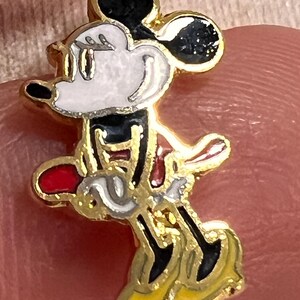 May include: A gold-toned metal charm featuring a colorful enamel inlay of a cartoon mouse character. The character has a white face, black ears, a red skirt, and yellow shoes, depicted in a side profile pose with a gold-colored metal border.