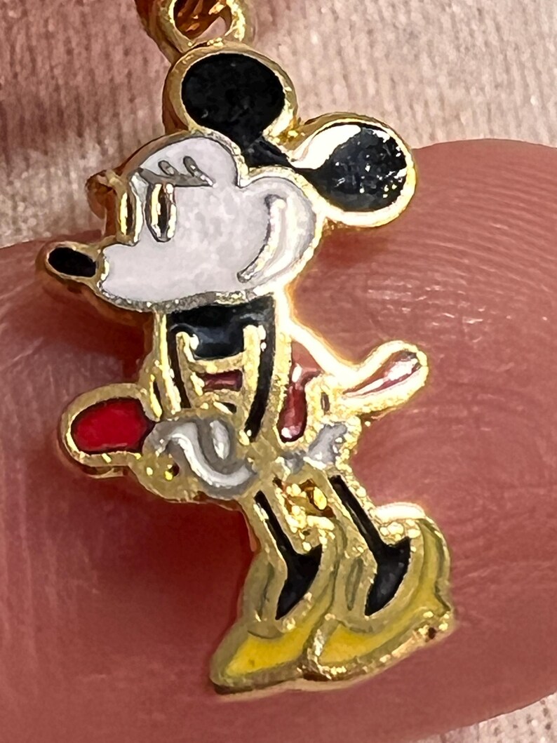 May include: A gold-toned metal charm featuring a colorful enamel inlay of a cartoon mouse character. The character has a white face, black ears, a red skirt, and yellow shoes, depicted in a side profile pose with a gold-colored metal border.