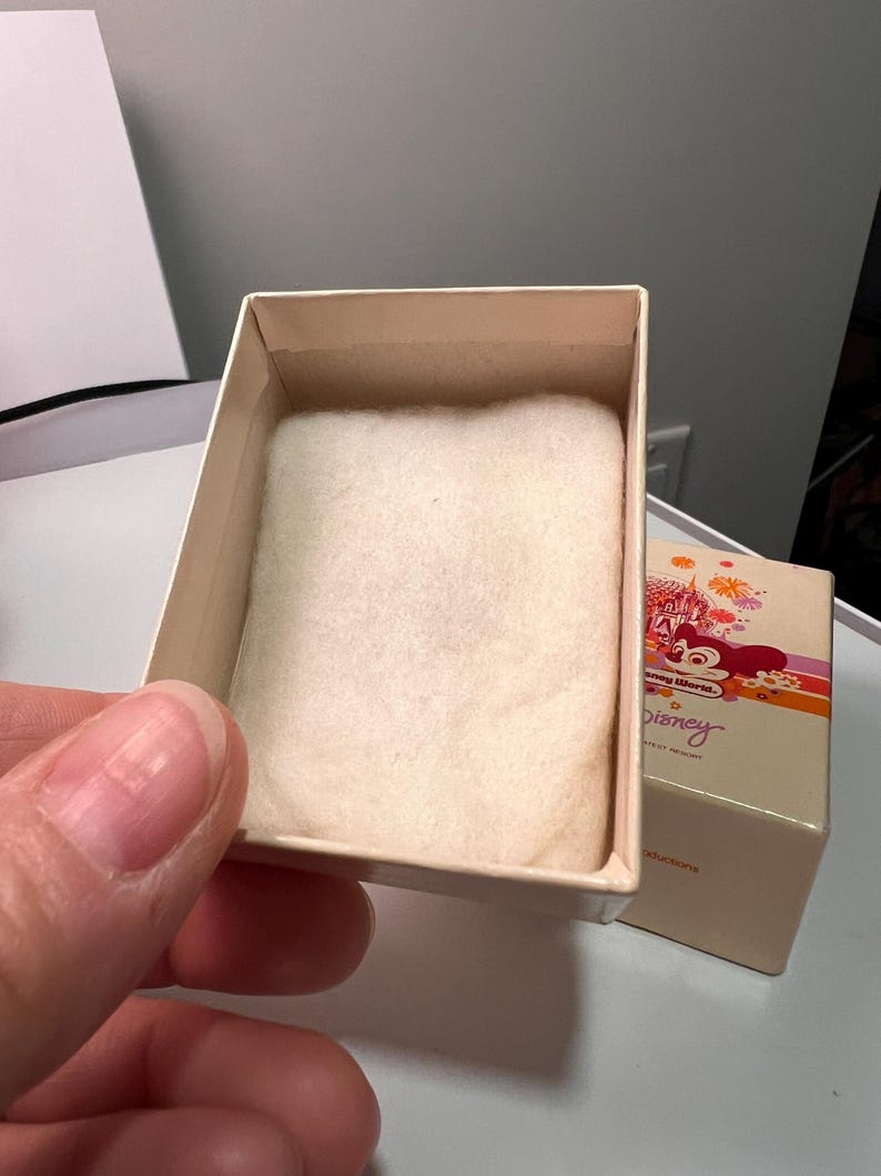 May include: A small, rectangular, cream-colored cardboard box with a soft, white, rectangular fabric insert inside. The box lid features a colorful graphic of a castle, fireworks, and the text Disney World and Disney.
