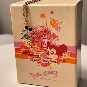 May include: A gold-toned metal necklace featuring a small Minnie Mouse charm, resting against a rectangular cream-colored box. The box displays a colorful graphic of a castle, fireworks, and the text Walt Disney World and Together at Disney.