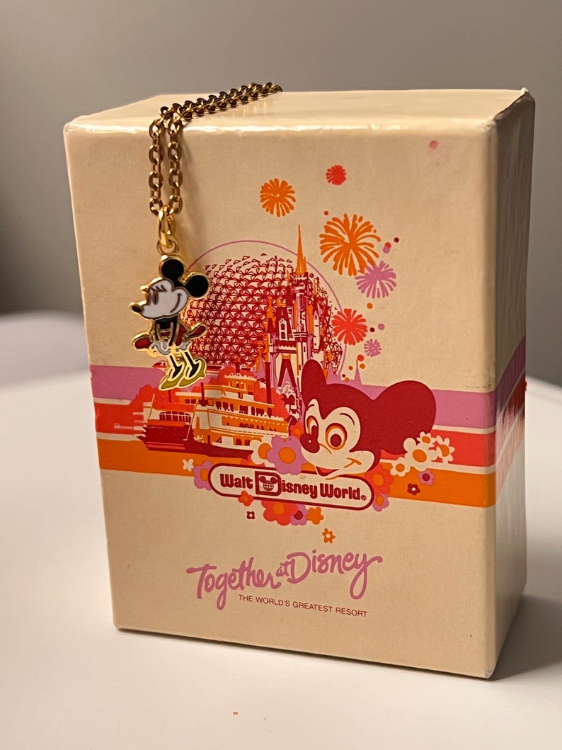 May include: A gold-toned metal necklace featuring a small Minnie Mouse charm, resting against a rectangular cream-colored box. The box displays a colorful graphic of a castle, fireworks, and the text Walt Disney World and Together at Disney.