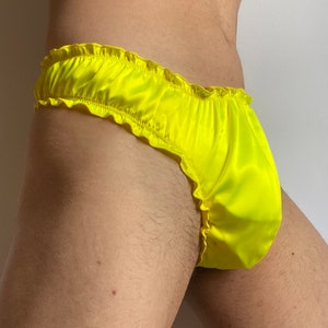 Pode incluir: Roupa interior de cetim amarelo néon brilhante com cintura elástica com folhos e aberturas para as pernas. O tecido tem um acabamento liso e brilhante com uma textura franzida ao longo das bordas, proporcionando um ajuste justo ao corpo.