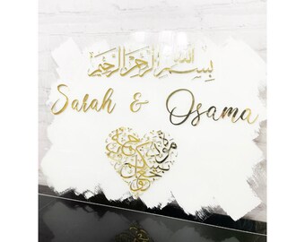 May include: White acrylic sign with gold lettering. The sign features the names "Sarah & Osama" and a heart-shaped design with Arabic calligraphy. The background is a white brushstroke.