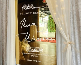 May include: A gold-framed mirror sign with white calligraphy and Arabic script, announcing "Welcome to the Nikkah" and the names "Ikram and Warsame." The date "February 18, 2024" is also displayed. The sign is draped with white fabric.