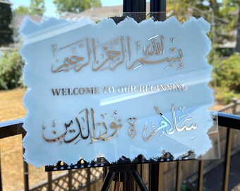 May include: A light blue acrylic sign with silver lettering. The sign says "Welcome to our beginning" and features Arabic calligraphy. The sign is on a black easel.
