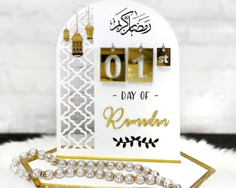 May include: White wooden Ramadan countdown calendar with gold accents. The calendar features a crescent moon, lanterns, and the Arabic word for Ramadan. The calendar is set to the 1st day of Ramadan. A white prayer bead necklace is on the base of the calendar.