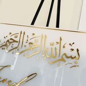 May include: White square sign with gold Arabic calligraphy that reads "Bismillah al-Rahman al-Rahim" and gold cursive writing that reads "Blessed".