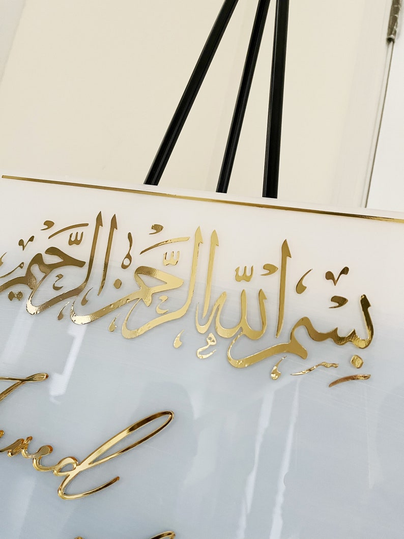 May include: White square sign with gold Arabic calligraphy that reads "Bismillah al-Rahman al-Rahim" and gold cursive writing that reads "Blessed".