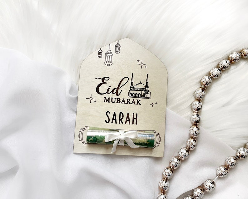 Puede incluir: Un letrero de madera con el texto "Eid Mubarak" y una ilustraci&oacute;n de una mezquita. El letrero tambi&eacute;n tiene el nombre "Sarah" escrito en &eacute;l. Un regalo enrollado se coloca delante del letrero.