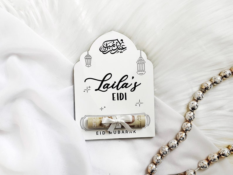 May include: White card with a black arch design and the words "Laila's Eidi" in a decorative font. The card has a rolled up bill of money with a white ribbon tied around it. The card also has the words "Eid Mubarak" printed on it.