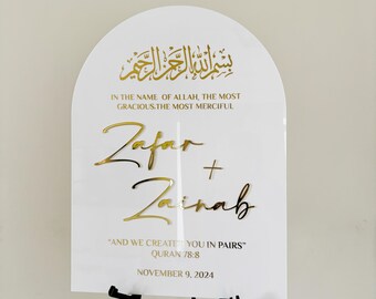 May include: A white sign with gold lettering announcing a wedding. The sign reads "Bismillah ar-Rahman ar-Rahim" in Arabic script, followed by "In the name of Allah, the most gracious, the most merciful." The names "Zafar" and "Zainab" are written in cursive script, followed by the quote "And we created you in pairs" from the Quran, verse 78:8. The date "November 9, 2024" is printed at the bottom.
