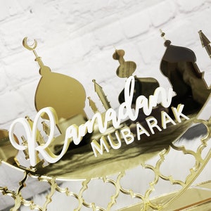 May include: Gold mirrored Ramadan Mubarak sign with a crescent moon and mosque silhouettes. The sign is decorated with a gold geometric pattern.