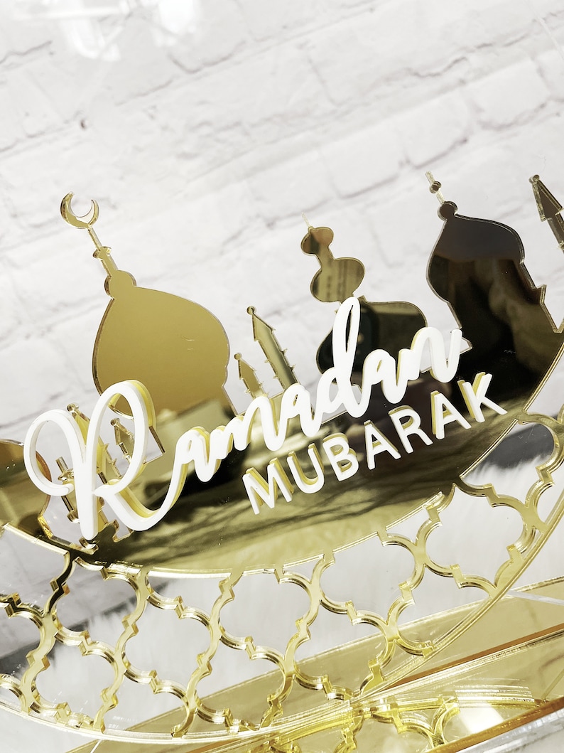 May include: Gold mirrored Ramadan Mubarak sign with a crescent moon and mosque silhouettes. The sign is decorated with a gold geometric pattern.