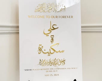 May include: A white sign with gold lettering and Arabic calligraphy, displayed on a black easel. The sign reads "WELCOME TO OUR FOREVER" and includes a date, May 25, 2024, and a quote from the Quran.