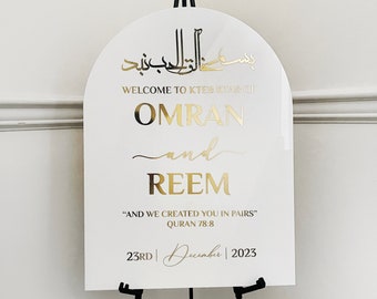 May include: White arch-shaped sign with gold lettering announcing the wedding of Omran and Reem. The sign includes the Arabic phrase "Bismillah al-Rahman al-Rahim" and the Quran verse 78:8, "And We created you in pairs." The date of the wedding is December 23rd, 2023.