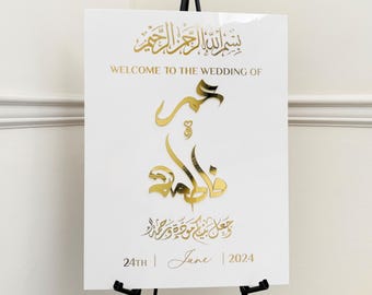 May include: White wedding sign with gold calligraphy that reads "بسم الله الرحمن الرحيم" and "WELCOME TO THE WEDDING OF" in English. The Arabic calligraphy reads "جعل بن مودة ورحمة". The date is "24TH June 2024".