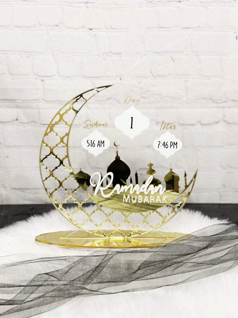 May include: Gold Ramadan Mubarak countdown stand with a crescent moon design. The stand features a day counter, Suhoor time, and Iftar time. The text "Ramadan Mubarak" is displayed on the stand.