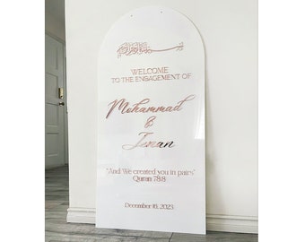 May include: White acrylic arch sign with a rose gold inscription announcing the engagement of Mohammad and Jenan. The sign includes the Arabic phrase "Bismillah" and a Quran verse: "And We created you in pairs" (Quran 78:8). The date December 16, 2023 is also included.