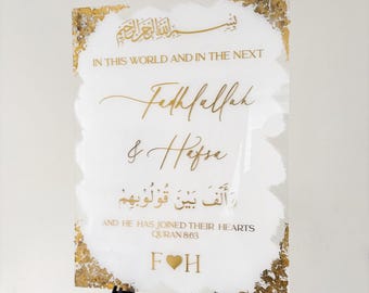 May include: A white sign with gold accents and calligraphy announcing the wedding of Fathfallah and Hafse. The sign includes a Quran verse in Arabic script and the text "And He has joined their hearts, Quran 863".