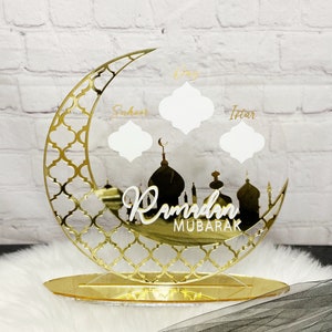 May include: Gold Ramadan Mubarak sign with a crescent moon and mosque silhouette. The sign has three blank spaces for writing in the names of the days of Ramadan: Suhoor, Iftar, and Day.